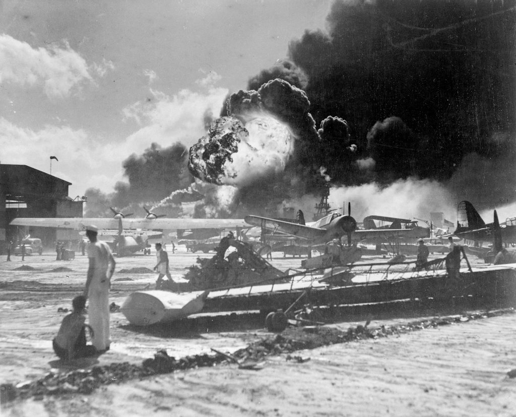 Sailors among wrecked planes at the Ford Island seaplane base as USS Shaw explodes during the Pearl Harbor attack